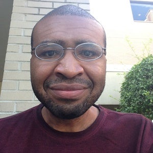 Justin is a black man smiling and wearing round glasses and a maroon shirt. There is a brick wall, a tree, and a window behind him