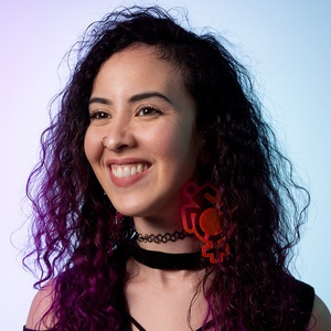 Genevieve Ramos is a smiling Mexican-American woman with curly shoulder length hair that is dark with red dye at the ends. She is wearing oversized hot pink earrings and a black top